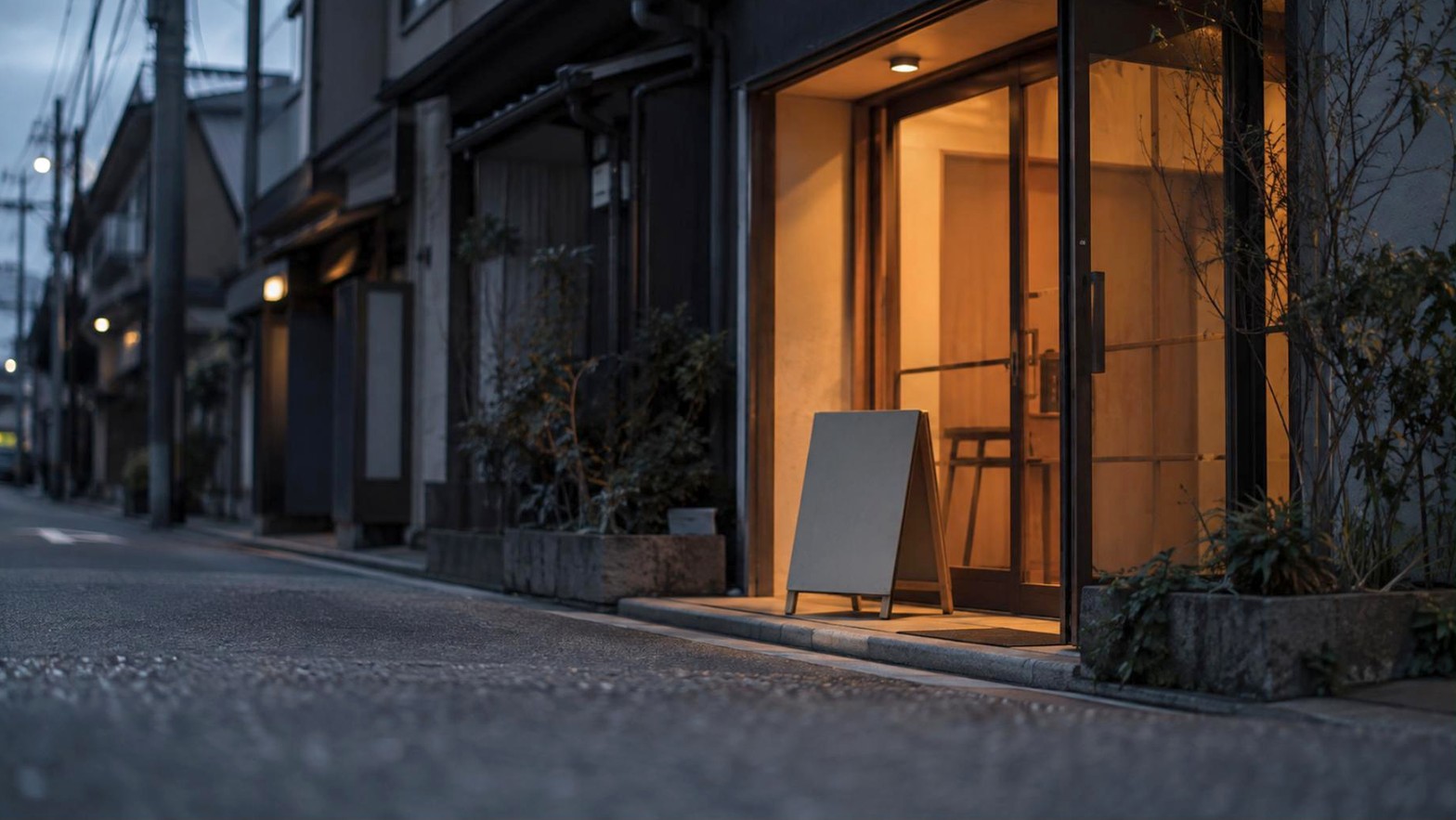 夕方の通りに面した小さな店の入り口。ガラス戸の向こうに灯りがともり、手前の歩道に立て看板が出ている