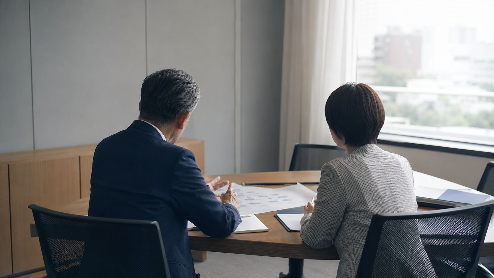 窓際の机に並んで座り、資料を見ながら話す2人